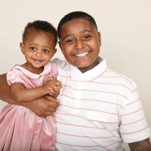 Proud siblings, big brother gives his little sister a cuddle, neutral backdrop and pinks